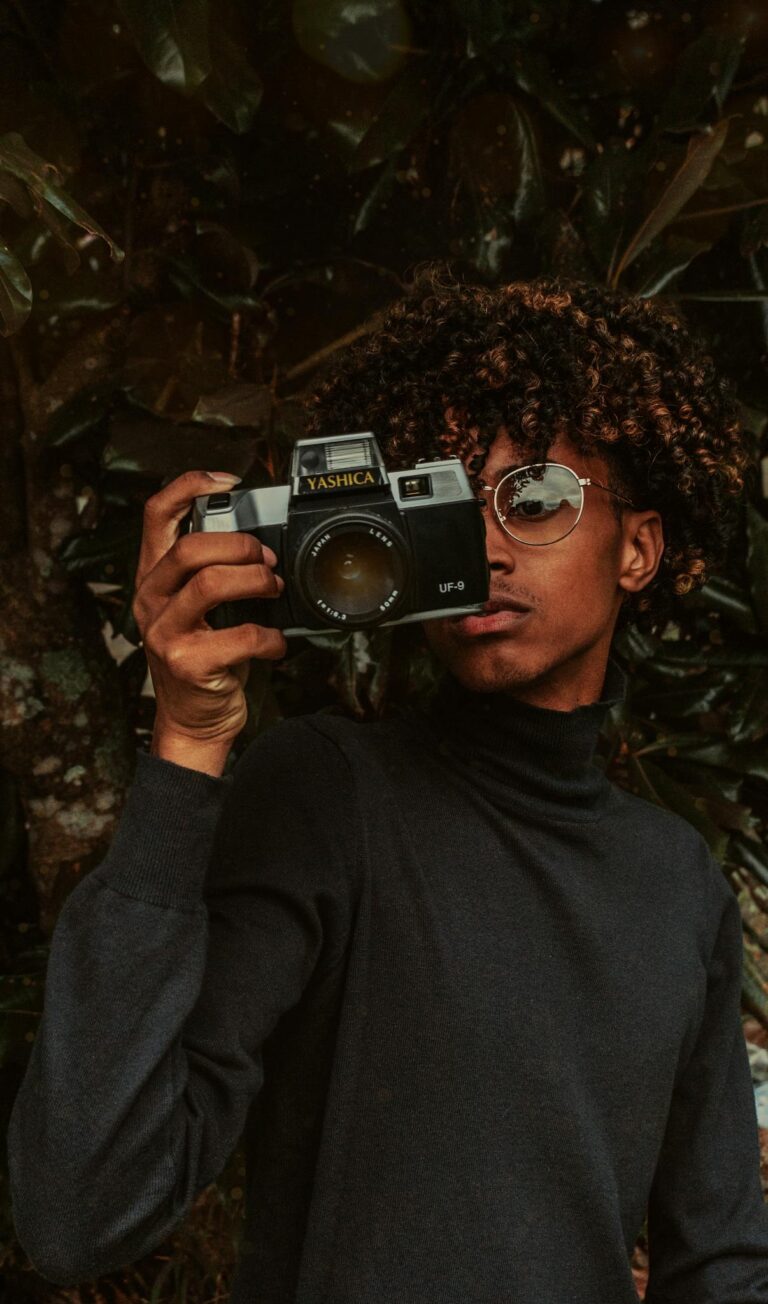 Stylish African American young male millennial in turtleneck and eyeglasses taking picture on vintage photo camera while standing near green tree in park