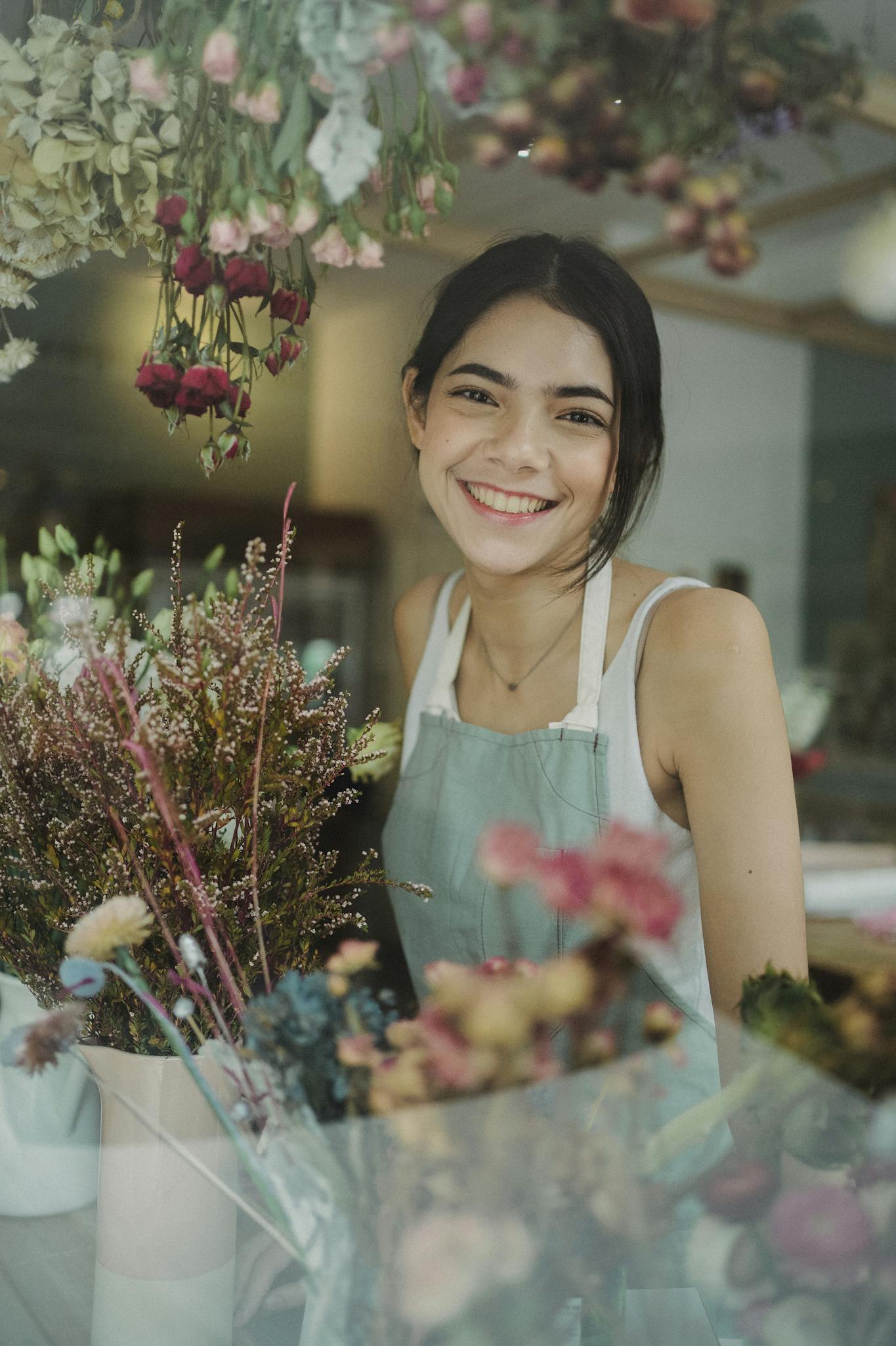 Through window of charming female worker in apron standing among flowers in store smiling at camera